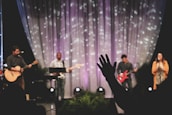 A musical performance on stage features a group of musicians, each with an instrument. The backdrop includes softly lit drapery with scattered light patterns. A person in the foreground raises a hand in what seems to be an expression of involvement or enjoyment.