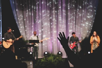 A musical performance on stage features a group of musicians, each with an instrument. The backdrop includes softly lit drapery with scattered light patterns. A person in the foreground raises a hand in what seems to be an expression of involvement or enjoyment.