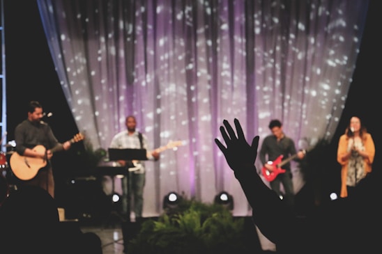 A musical performance on stage features a group of musicians, each with an instrument. The backdrop includes softly lit drapery with scattered light patterns. A person in the foreground raises a hand in what seems to be an expression of involvement or enjoyment.