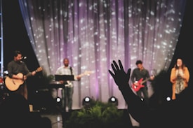 A musical performance on stage features a group of musicians, each with an instrument. The backdrop includes softly lit drapery with scattered light patterns. A person in the foreground raises a hand in what seems to be an expression of involvement or enjoyment.