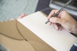 A hand holding a black pen poised above an open brown notebook with lined pages. The background is softly blurred, focusing attention on the writing task.