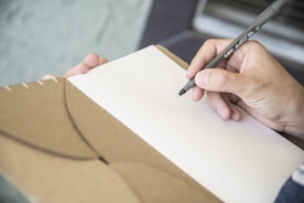 A hand holding a black pen poised above an open brown notebook with lined pages. The background is softly blurred, focusing attention on the writing task.