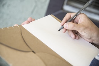 A hand holding a black pen poised above an open brown notebook with lined pages. The background is softly blurred, focusing attention on the writing task.