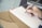 A hand holding a black pen poised above an open brown notebook with lined pages. The background is softly blurred, focusing attention on the writing task.