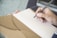 A hand holding a black pen poised above an open brown notebook with lined pages. The background is softly blurred, focusing attention on the writing task.