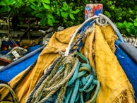 A close-up of a bundle of ropes intertwined and placed on a weathered yellow and blue fabric or tarp, set against a backdrop of green leaves and a wooden structure.