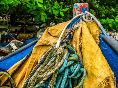 A close-up of a bundle of ropes intertwined and placed on a weathered yellow and blue fabric or tarp, set against a backdrop of green leaves and a wooden structure.
