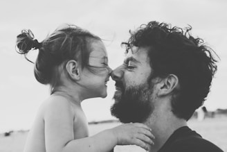 A black-and-white portrait captures an intimate moment between a man and a young child, both facing each other closely with gentle smiles. The child is holding the man's shoulder, while both exhibit expressions of affection and joy.