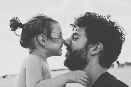 A black-and-white portrait captures an intimate moment between a man and a young child, both facing each other closely with gentle smiles. The child is holding the man's shoulder, while both exhibit expressions of affection and joy.
