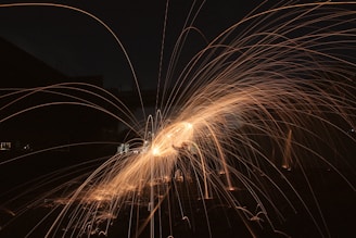 Long exposure photograph capturing bright, orange sparks flying in arcs against a dark background, creating a dynamic and artistic pattern.