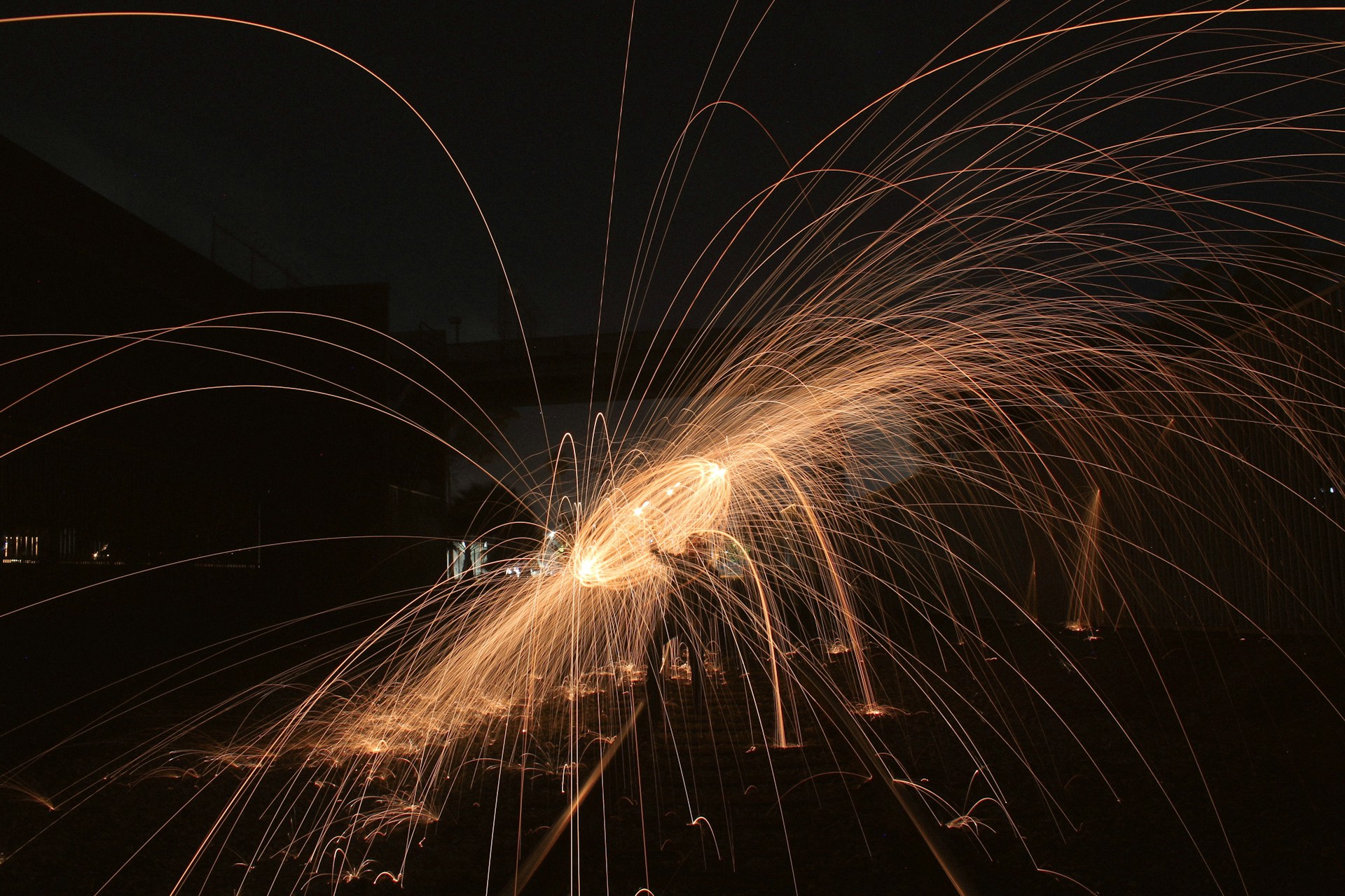 Long exposure photograph capturing bright, orange sparks flying in arcs against a dark background, creating a dynamic and artistic pattern.