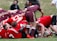 A group of rugby players are engaged in a scrum on a grassy field. The players are wearing maroon and red uniforms with white stripes. A rugby ball is visible on the ground, partially held by one player. Spectators can be seen blurred in the background.