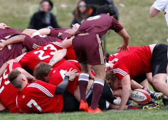 A group of rugby players are engaged in a scrum on a grassy field. The players are wearing maroon and red uniforms with white stripes. A rugby ball is visible on the ground, partially held by one player. Spectators can be seen blurred in the background.