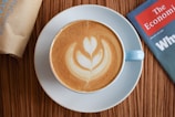 A cup of coffee with latte art in the shape of a leaf is placed on a white saucer. The coffee is in a light blue cup. A magazine titled 'The Economist' is partially visible on the right side, placed on a wooden table, and a brown paper bag is on the left.