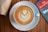 A cup of coffee with latte art in the shape of a leaf is placed on a white saucer. The coffee is in a light blue cup. A magazine titled 'The Economist' is partially visible on the right side, placed on a wooden table, and a brown paper bag is on the left.