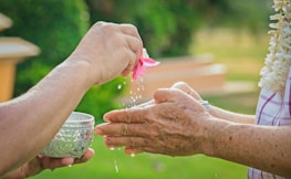 Two hands are seen holding small, ornate silver bowls. Water is being poured from one bowl to another, with the addition of a pink flower, creating droplets. The background suggests a natural setting with greenery.