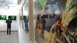 A person stands in an art gallery observing framed photographs on the wall. One of the photographs shows a portrait of a woman engaged in an outdoor activity. The gallery has a clean, minimalist design with soft lighting and a reflective surface to the right capturing part of the scene.