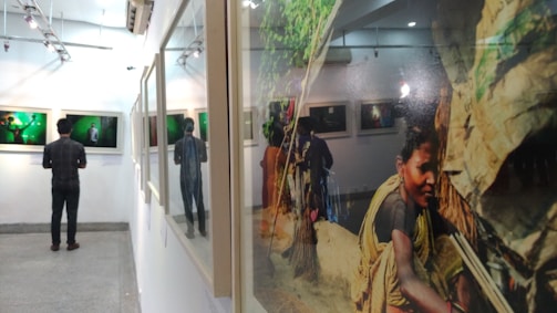 A person stands in an art gallery observing framed photographs on the wall. One of the photographs shows a portrait of a woman engaged in an outdoor activity. The gallery has a clean, minimalist design with soft lighting and a reflective surface to the right capturing part of the scene.