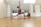 A person is performing a yoga pose on a red mat in a spacious, well-lit room with light wooden flooring and white walls. The individual is wearing a red top and gray leggings and is in a warrior pose, balancing on one leg with arms reaching upwards. The room has a minimalist design with open windows and a stack of colorful yoga mats in the corner.