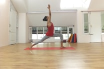 A person is performing a yoga pose on a red mat in a spacious, well-lit room with light wooden flooring and white walls. The individual is wearing a red top and gray leggings and is in a warrior pose, balancing on one leg with arms reaching upwards. The room has a minimalist design with open windows and a stack of colorful yoga mats in the corner.