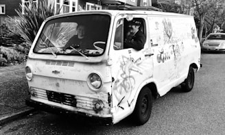A vintage van covered in graffiti and drawings is parked on the street. Two people are visible inside the vehicle, with the driver looking out the window and appearing to wave or perform a gesture. The van has a rugged, worn appearance, adding character to its presence on the road. Surrounding the van are residential buildings and vegetation, contributing to an urban yet laid-back environment.