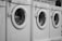 A row of white front-loading washing machines is numbered sequentially. Tags with keys hang on each machine's handle, indicating service numbers. Towels or clothes are visible inside one of the machines.