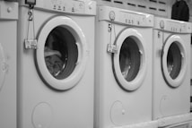 A row of white front-loading washing machines is numbered sequentially. Tags with keys hang on each machine's handle, indicating service numbers. Towels or clothes are visible inside one of the machines.