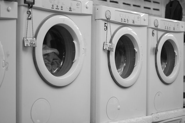 A row of white front-loading washing machines is numbered sequentially. Tags with keys hang on each machine's handle, indicating service numbers. Towels or clothes are visible inside one of the machines.