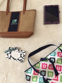 A beige tote bag with brown leather accents and the word 'HOME' embroidered on it is positioned next to a tablet in a purple case. Below, a small decorative card with a wrist strap featuring a black leaf pattern is seen beside a colorful bag with geometric designs in vibrant colors.