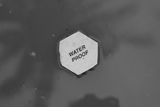 A hexagonal piece of material floating on water with the text 'WATER PROOF' written on it. The background is a monochromatic gray, suggesting a watery surface with ripples around the object.