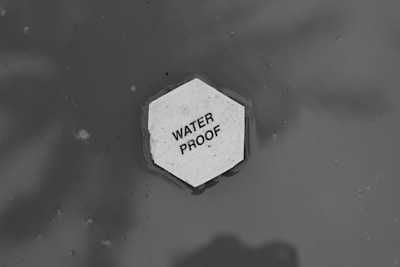 A hexagonal piece of material floating on water with the text 'WATER PROOF' written on it. The background is a monochromatic gray, suggesting a watery surface with ripples around the object.