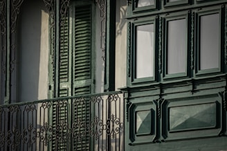 A detailed ornamental iron balcony railing with intricate patterns stands in front of a building facade featuring closed green wooden shutters and large, decorative green-paneled windows.