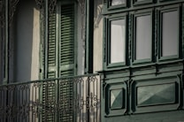 A detailed ornamental iron balcony railing with intricate patterns stands in front of a building facade featuring closed green wooden shutters and large, decorative green-paneled windows.