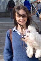 A woman smiles while holding a small fluffy dog adorned with a red bow on its ear. Her hair is slightly windswept, and she wears a blue sweater over a white shirt. The background features a set of stairs with several people walking.