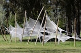 White netting structures are artistically draped between tall wooden poles in an outdoor setting. The scene is surrounded by lush green grass and dense trees, creating a natural, serene environment.