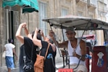 A person in a tank top and cap is seated in a bicycle rickshaw parked on a street lined with historic buildings. Two tourists are photographing nearby architecture, focusing upwards at the old stone facade. A pedestrian in a denim skirt and white top walks on the sidewalk under a green awning.
