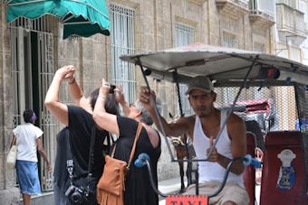 A person in a tank top and cap is seated in a bicycle rickshaw parked on a street lined with historic buildings. Two tourists are photographing nearby architecture, focusing upwards at the old stone facade. A pedestrian in a denim skirt and white top walks on the sidewalk under a green awning.