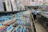 A record store aisle filled with vinyl records organized by categories, each marked with blue tab labels. The shelves are packed tightly with records, and in the background, a person is browsing through the collection. Posters and album artwork decorate the walls above the shelves.