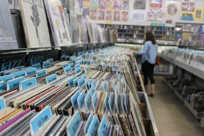 A record store aisle filled with vinyl records organized by categories, each marked with blue tab labels. The shelves are packed tightly with records, and in the background, a person is browsing through the collection. Posters and album artwork decorate the walls above the shelves.