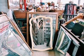 A cluttered space filled with various items like mirrors, frames, baskets, and books. A person is captured in the reflection of a uniquely shaped mirror in the center, wearing a long patterned dress and holding a camera. The surroundings suggest an antique or thrift shop atmosphere with a variety of objects on and around shelves.