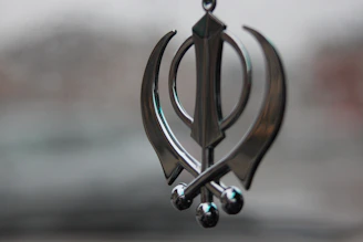 A metallic ornament shaped like a Khanda symbol, featuring a central double-edged sword flanked by two single-edged swords and a circular element in the background. The ornament hangs against a blurred, neutral background.