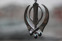 A metallic ornament shaped like a Khanda symbol, featuring a central double-edged sword flanked by two single-edged swords and a circular element in the background. The ornament hangs against a blurred, neutral background.