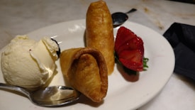A dessert plate features two pieces of fried pastry, a scoop of vanilla ice cream, and a sliced strawberry arranged aesthetically. A spoon is placed beside the ice cream and a black napkin is partially visible on the table.
