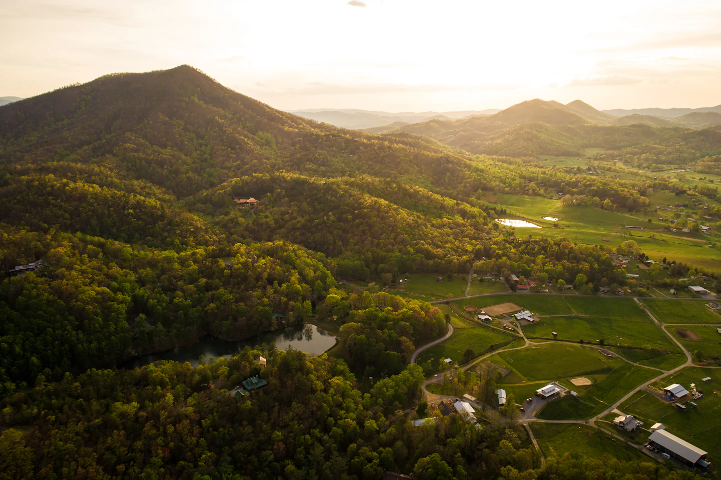 Farm buildings in a grassy valley below forested mountains.