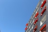 A modern building facade with windows featuring red awnings against a clear blue sky. The building's walls are grey with a grid-like pattern. The awnings create a contrast with the neutral tones of the structure, and a few flower pots can be seen on some windowsills.