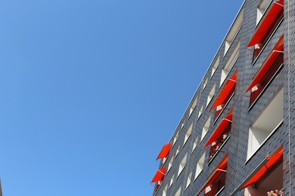 A modern building facade with windows featuring red awnings against a clear blue sky. The building's walls are grey with a grid-like pattern. The awnings create a contrast with the neutral tones of the structure, and a few flower pots can be seen on some windowsills.
