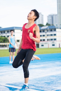 A young athlete in a red tank top and black leggings sprints on a blue track with a determined expression. The background shows a blurred person and city buildings, indicating a track event in a stadium.