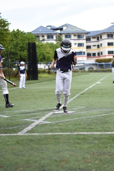 A baseball player wearing a helmet and uniform is stepping on home plate. Other players in similar uniforms are seen holding baseball bats on a grassy field. There is a background of trees and a building with blue rooftops.