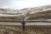 A person with hiking gear, including a red backpack, stands on rocky terrain, looking out over a snow-dappled landscape with mountains in the distance. A small stream or river flows across the foreground, winding between patches of snow and earth.