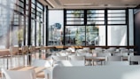 A spacious and modern cafeteria interior with multiple white tables and chairs. Large windows allow natural light to flood the area, providing a view of urban buildings and greenery outside. The setting is clean and organized, with wooden elements adding warmth to the space.